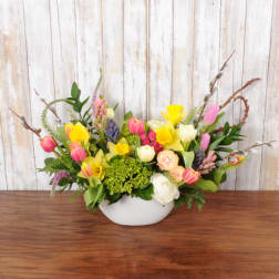 Low oval arrangement of pink tulips, yellow daffodils, roses, and mixed spring flowers in a white bowl