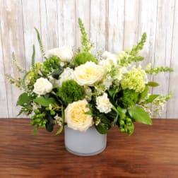 Low vase arrangement of cream roses, white flowers, and green accents in a frosted cylinder container.