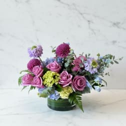 Low arrangement of mauve roses with purple mums and green and blue hydrangea in a glass vase
