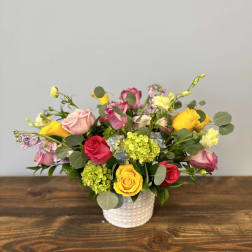 Low arrangement of mixed roses and other blooms in pink, yellow, and red in a white vase