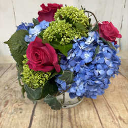 Low round arrangement of hot pink roses, green blooms, and blue hydrangeas in a clear glass vase