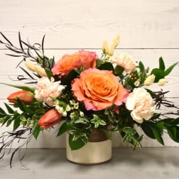 Low arrangement of peach roses, blush carnations, and orange tulips in a cream ceramic vase