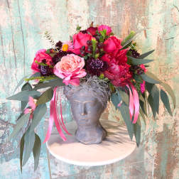 Pink and red floral arrangement in a gray bust vase with long ribbon streamers