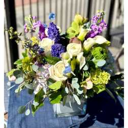Mixed arrangement of white roses and calla lilies with purple and blue blooms in a square glass vase