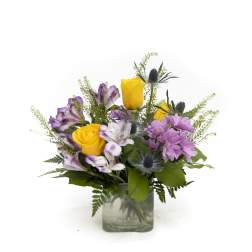 Mixed bouquet of yellow roses and purple flowers in a clear glass cube vase