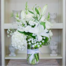 White lilies, roses, and hydrangea arranged in a clear glass vase tied with a white ribbon