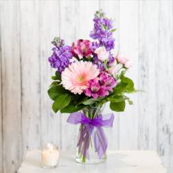 Mixed bouquet of pink and purple flowers in a clear vase with purple bow beside a lit candle.