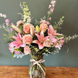 Tall bouquet of pink lilies, peach roses, and mixed blooms in a clear glass vase with a pink ribbon.