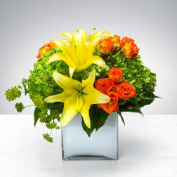 Low arrangement of yellow lilies and orange roses in a clear glass cube vase with bright green blooms.