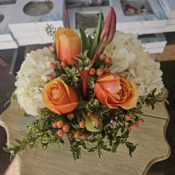 Compact arrangement of orange roses, tulip buds, white hydrangeas, and berries on a small table.