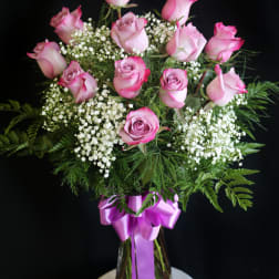 Pink roses in a glass vase with white filler flowers and a purple ribbon