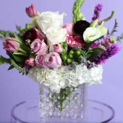 Mixed bouquet of roses, hydrangeas, and purple blooms in a clear vase