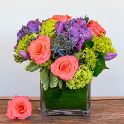 Short arrangement of coral roses, purple blooms, and green hydrangea in a square glass vase with a leaf wrap