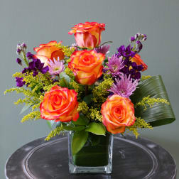Bright arrangement of orange roses and purple flowers in a glass cube vase with a folded green leaf