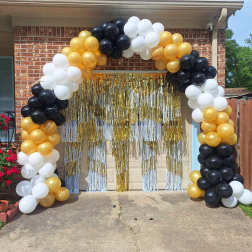 Black, white, and gold balloon arch framing a garage doorway with gold metallic fringe curtains.