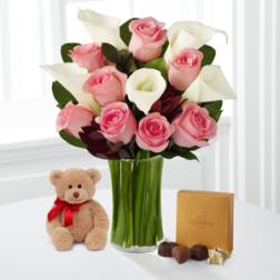 Pink roses and white calla lilies in a glass vase with a teddy bear and chocolates