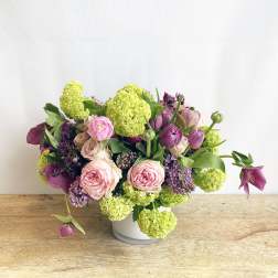 Low arrangement of pink roses, purple blooms, and green flower clusters in a white vase