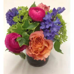 Short arrangement of peach rose, pink ranunculus, and purple flowers in a small glass vase
