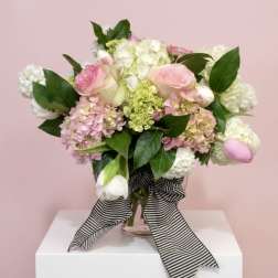 Pink and white roses, hydrangeas, and tulips in a glass vase with a striped ribbon