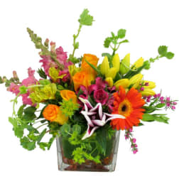 Bright mixed arrangement of orange roses, yellow lilies and assorted blooms in a square glass vase.