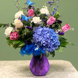 Mixed bouquet of blue hydrangea, pink roses, and white blooms in a purple glass vase