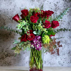 Red roses arranged in a clear glass vase with mixed greenery and purple hydrangea