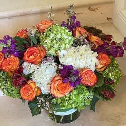 Low round arrangement of orange roses, green and white hydrangeas, and purple blooms in a glass vase