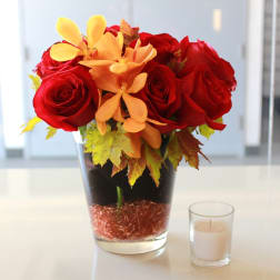 Red roses and orange flowers in a glass vase beside a small candle