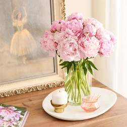 Pink peonies in a clear glass vase on a tray with a candle and small dish