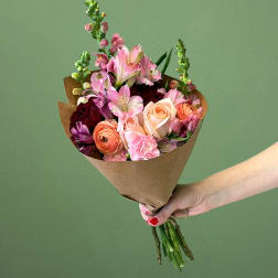 Hand-tied bouquet of pink and peach roses, ranunculus, and mixed blooms wrapped in brown paper.