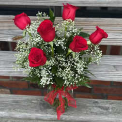 Glass vase of six red roses with white filler flowers and a red bow on a wooden bench