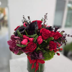 Red roses and dark purple flowers arranged in a glass vase with a red ribbon.