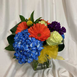 Colorful bouquet with blue hydrangea, orange gerbera daisies, and a yellow ribbon in a glass vase