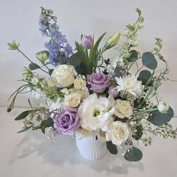 Pastel arrangement of white and lavender roses, lisianthus and delphinium in a white ribbed vase.