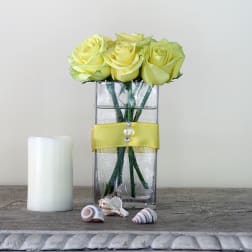 Yellow roses in a clear rectangular vase with a yellow ribbon and candle beside it