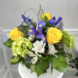 Compact arrangement of yellow roses, purple irises, green hydrangeas, and white alstroemeria in a glass vase