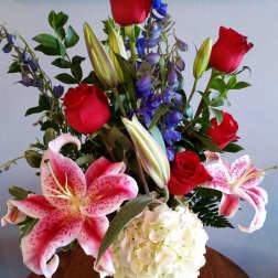 Mixed bouquet with red roses, pink lilies, blue delphinium, and white hydrangea in a lavender vase