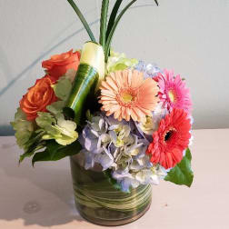 Compact arrangement of bright gerbera daisies, roses, and hydrangeas in a short glass vase with tall looping grass.