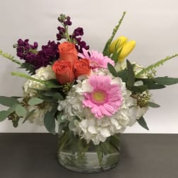 Low mixed arrangement of white hydrangeas, pink gerberas, orange roses, and yellow tulips in a glass vase