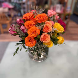 Mixed bouquet of orange, pink, and yellow roses with white filler flowers in a clear glass vase