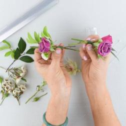 Hands arranging small purple roses with greenery on a white surface