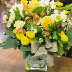 Yellow and white mixed flower arrangement with roses and daisies in a bee-themed glass cube vase with a burlap bow