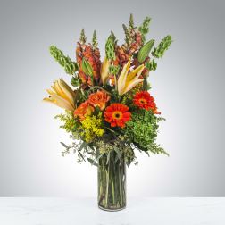 Tall arrangement of orange gerbera daisies, yellow lilies, and mixed blooms in a clear glass vase