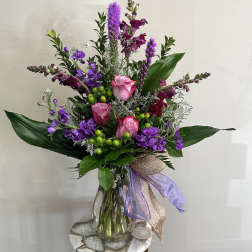 Tall arrangement of pink roses and purple flowers in a clear glass vase with decorative bow