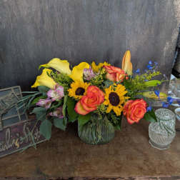 Mixed bouquet with roses, sunflowers, calla lilies, and blue accents in a glass vase