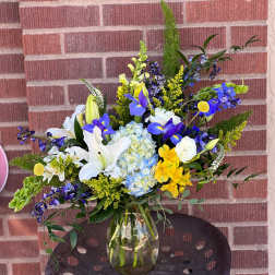 Mixed bouquet of white lilies, blue irises, and yellow flowers in a glass vase.