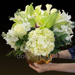 Low arrangement of white and pale green hydrangeas, roses, lilies, and an orchid in an amber glass cube vase.