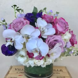 Round arrangement of white orchids, hydrangeas, and pink and purple roses in a glass vase.