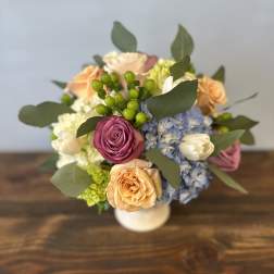 Compact arrangement of peach and mauve roses with blue hydrangeas in a white vase