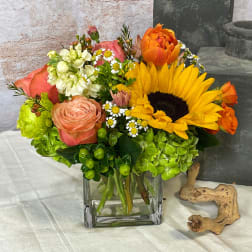 Mixed bouquet with a sunflower, roses, and orange blooms in a square glass vase